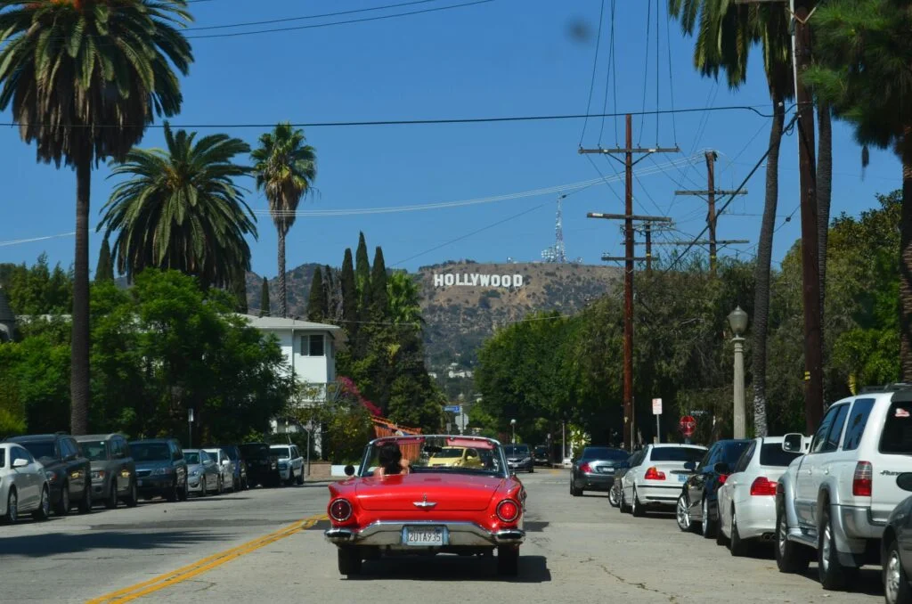 A view down a street lined with parked cars leading towards the iconic Hollywood sign on the hillside under a clear blue sky. A red vintage convertible car is driving towards the camera, and lush green trees can be seen on both sides of the street. The scene captures a quintessential Los Angeles vibe with its combination of urban environment and famous landmark.