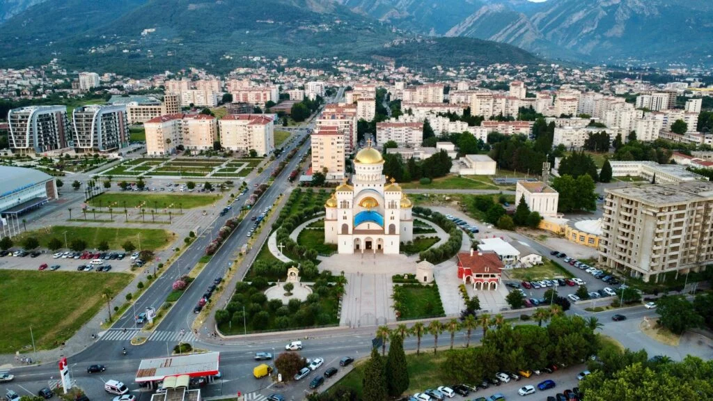 Aerial view of Bar, Montenegro, featuring a golden-domed church at the center, surrounded by pathways and greenery. The cityscape includes residential buildings against a mountainous backdrop.