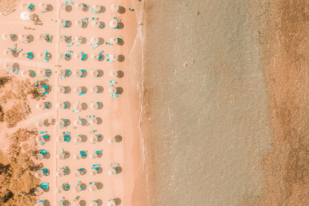 Aerial view of a sandy beach in Heraklion with clear turquoise waters on one side and a beachfront lined with colorful umbrellas and sun loungers on the other.