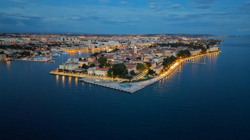 Aerial view of Zadar, Croatia during twilight, showcasing the historic city peninsula surrounded by the calm Adriatic Sea with lights beginning to illuminate the streets and buildings