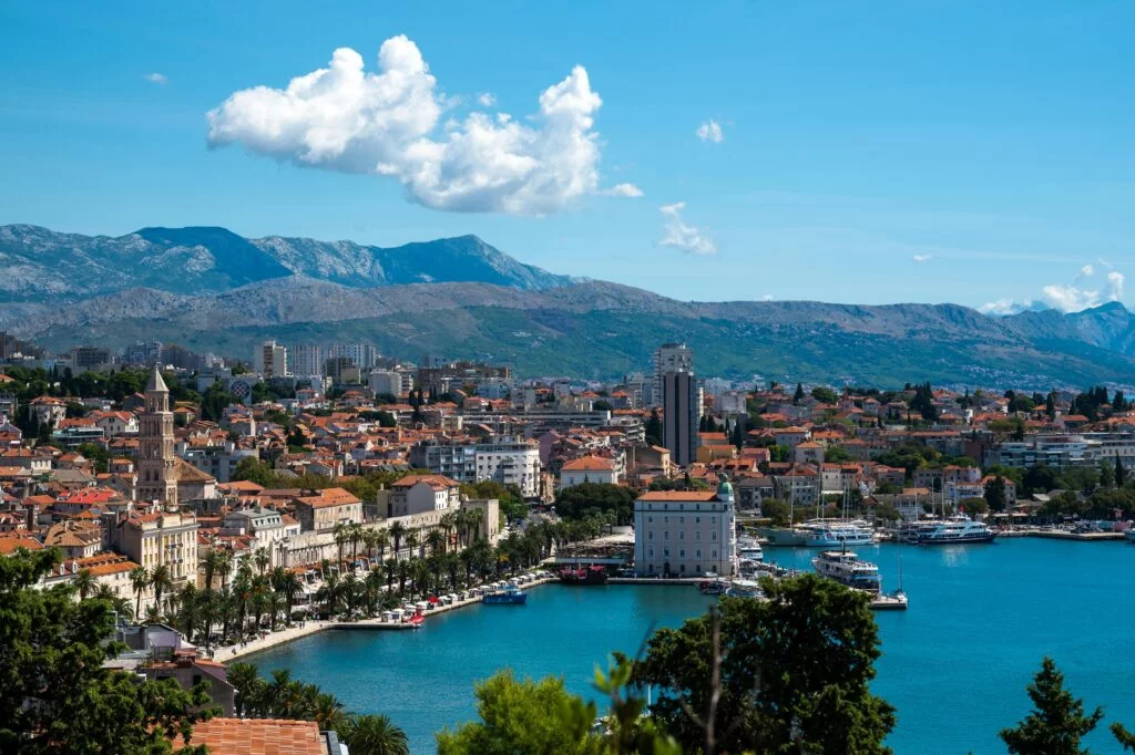 A panoramic view of Split, Croatia, with a clear blue sky. The cityscape includes various buildings with red-tiled roofs, nestled between the bright blue sea on the right and rolling hills in the background. Several boats are docked in the harbor, and lush greenery is interspersed throughout the city. A few scattered clouds float above, casting shadows on the mountainous terrain behind the urban area.