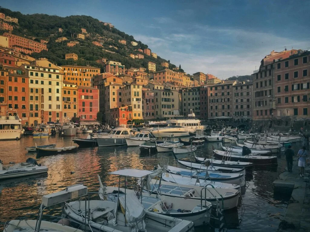 A scenic view of a harbor with numerous boats moored in calm waters. The background features a hillside densely packed with colorful buildings under a clear sky with soft clouds, reflecting the warm glow of sunset or sunrise. The architecture and the arrangement of the boats create a picturesque and tranquil maritime setting.