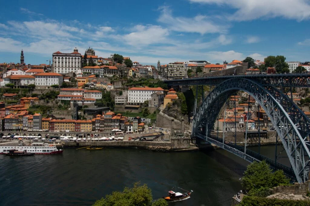 Panoramic view of Porto, Portugal, featuring the iconic Dom Luís I Bridge spanning the Douro River. The bridge’s double-deck metal arch is visible, with traditional Portuguese buildings ascending a hill in the background. A tour boat and clear sky complete the scene.