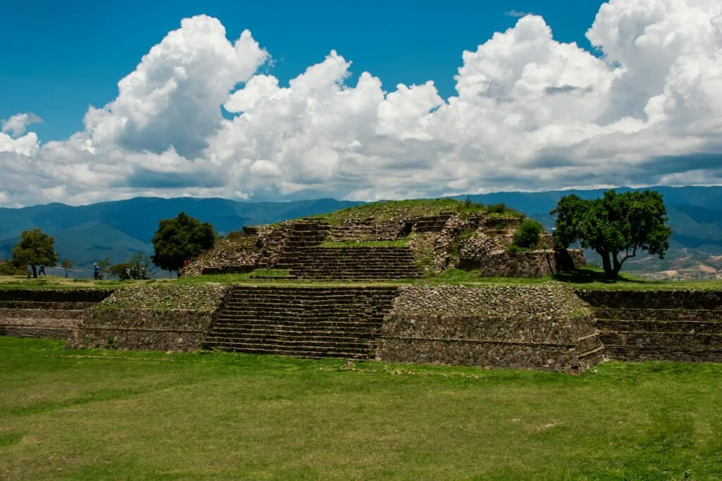 Monte Alban, Oaxaca, Mexico Image of Monte Albán, an ancient Zapotec city in Oaxaca, Mexico, showcasing a large pyramid-like structure with staircases amidst green grass, under a bright blue sky dotted with fluffy white clouds. In the background, rolling hills and mountains stretch across the horizon.