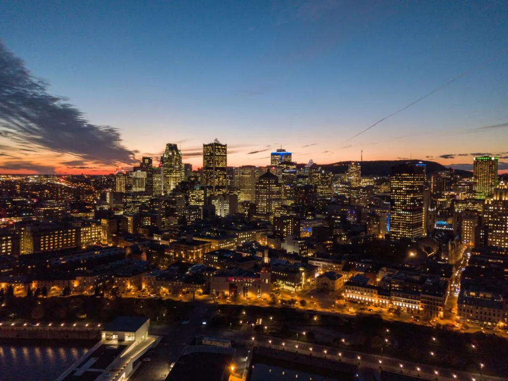 Aerial view of Montreal at dusk, showcasing the city’s skyline with illuminated buildings against a twilight sky with gradients of blue to orange. The urban landscape is dotted with lights, and the horizon is marked by the silhouettes of skyscrapers.