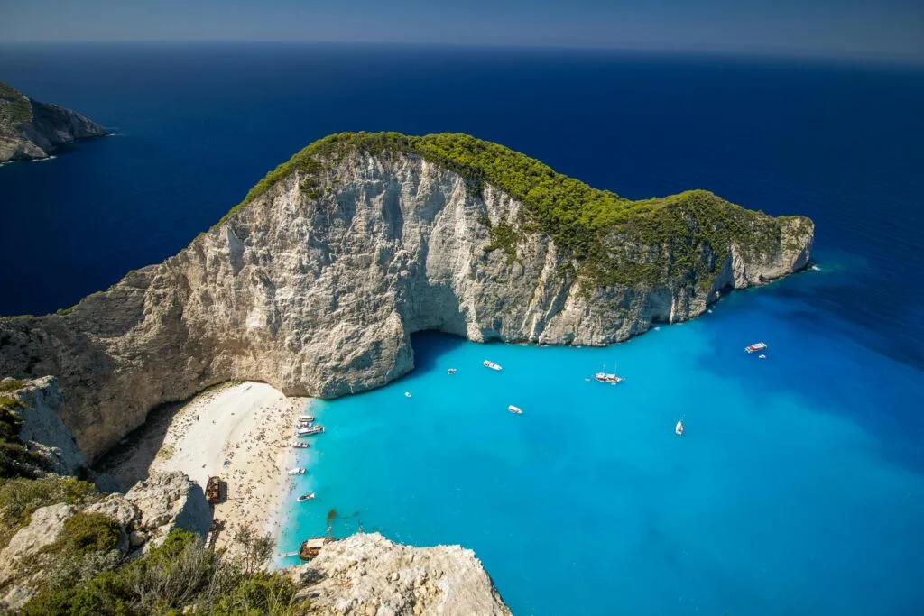 Aerial view of Navagio Beach, also known as Shipwreck Beach, in Zante, Greece. The image shows a large cove with steep white cliffs surrounding a small white sandy beach. The turquoise waters of the Ionian Sea are clear, allowing for the visibility of the sandy bottom near the shore. Several boats are floating on the water near the beach. A shipwreck is partially buried in the sand on one side of the beach, which is a notable feature of this popular tourist destination.