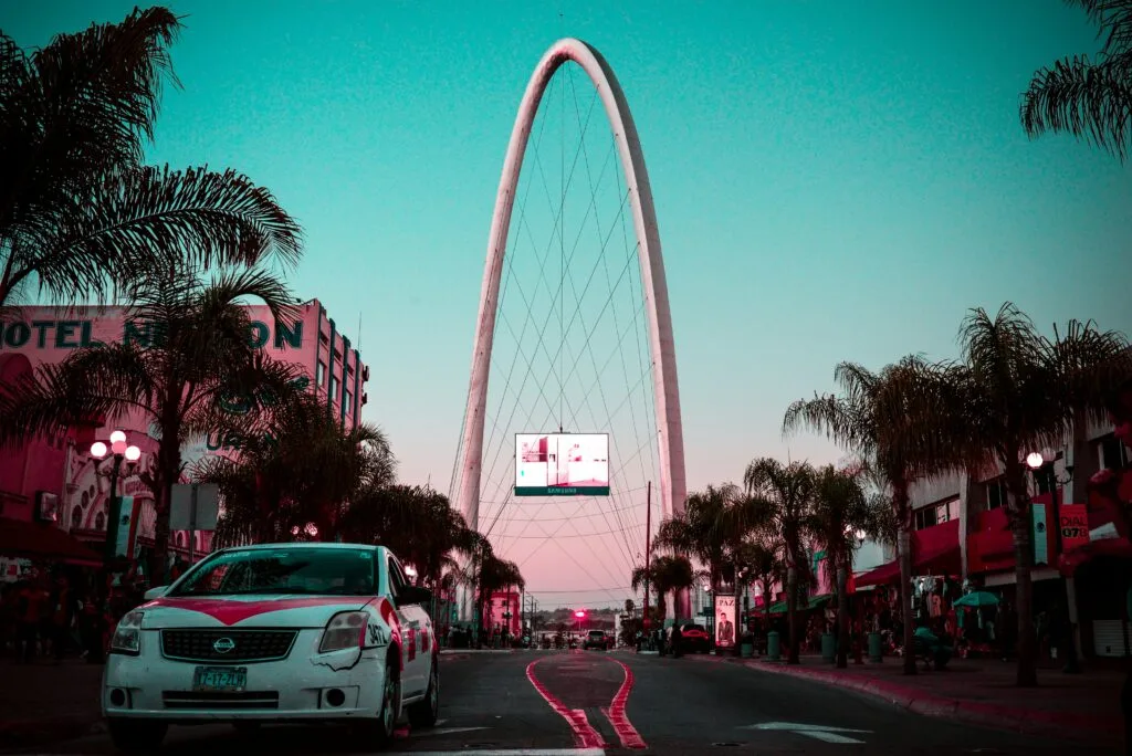 Urban street scene in Tijuana, Mexico, featuring the iconic arch-shaped Monumental Arch structure in the background, with a large digital screen displaying ads near its base. Palm trees line the street, creating a tropical feel, while a white taxi with a red design is parked in the foreground. Buildings with vibrant colors and signage, including one for 'Hotel Nelson,' add to the lively atmosphere. The sky has a surreal pink and teal gradient, enhancing the image's dramatic and vibrant look